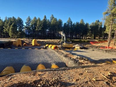 Construction site with excavator, yellow plastic drainage modules, gravel base, and trees in the background under a clear blue sky.