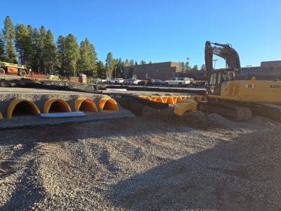 Construction site with a yellow excavator and multiple large yellow drainage pipes partially buried in gravel, with vehicles and trees in the background.