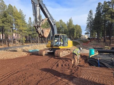 A construction worker levels dirt near a large yellow excavator at a job site surrounded by trees.