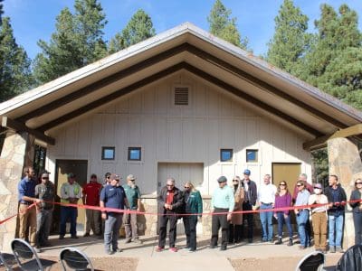 A group of people stands in front of a building during a ribbon-cutting ceremony, with chairs set up in front and trees in the background.