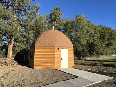 A small, dome-shaped cabin with a log-style exterior and a single white door, surrounded by trees and a grassy lawn under a clear blue sky.