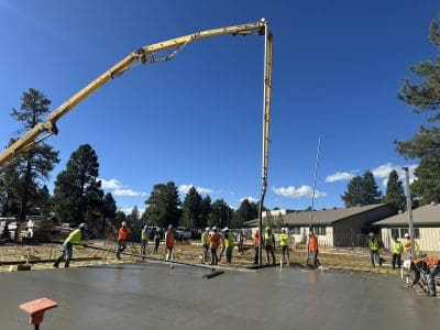 Construction workers in safety vests pour and smooth concrete on a building site using a large concrete pump, with trees and buildings in the background.
