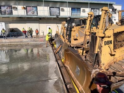 Construction workers operate bulldozers and smooth wet concrete at a building site with multiple workers and equipment visible.