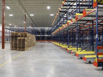Large warehouse interior with empty metal shelving units on the right and stacked wooden pallets on the left; concrete floor and bright lighting.