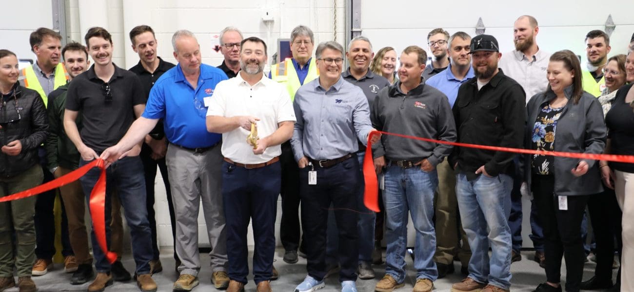 A group of people stand together at a Joy Baking Group Ribbon Cutting ceremony inside a building, with two men in the front holding large scissors and a piece of ribbon.
