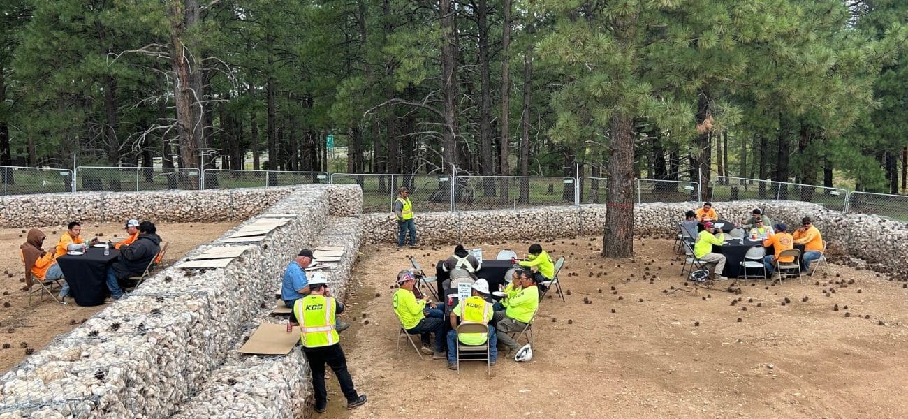Several groups of people in safety vests and orange shirts sit at tables outdoors among rock-filled barriers, surrounded by pine trees, as Kinney celebrates Earth Day—Building Better Together for a Greener Tomorrow.