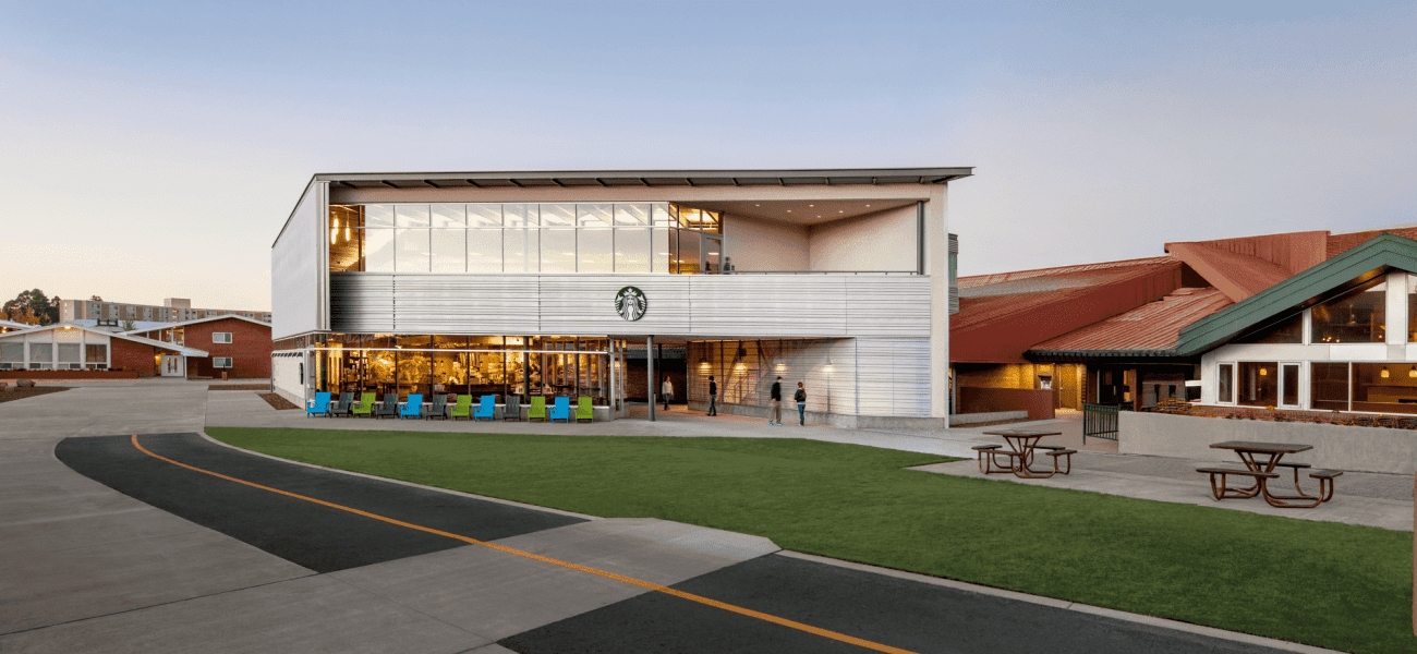 A modern two-story Starbucks building with large glass windows, outdoor seating, and a green lawn under a clear sky.