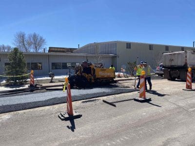 Construction workers operate machinery and lay asphalt on a road in front of a commercial building, with traffic barriers set up around the work area.