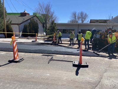 Construction workers repaving a section of road with asphalt, surrounded by caution tape and traffic cones on a sunny day.