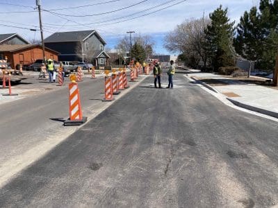 Road construction site with traffic barriers, workers in safety vests, and machinery on a residential street under a clear sky.