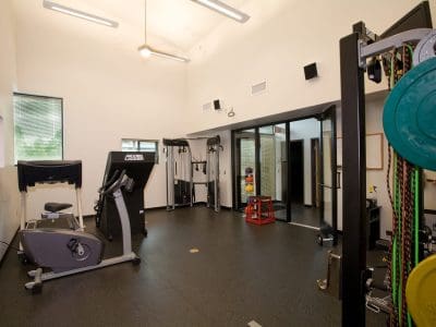 A modern gym room with exercise equipment, including a stationary bike, weight machine, resistance bands, and a rack of colorful weights against a white wall.