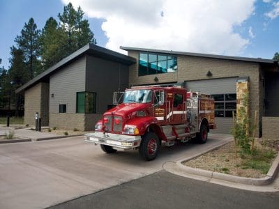 A red fire truck is parked in front of a modern fire station building surrounded by trees, under a partly cloudy sky.