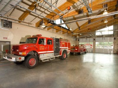 Two red fire trucks are parked inside a spacious, modern fire station garage with high ceilings and exposed beams.