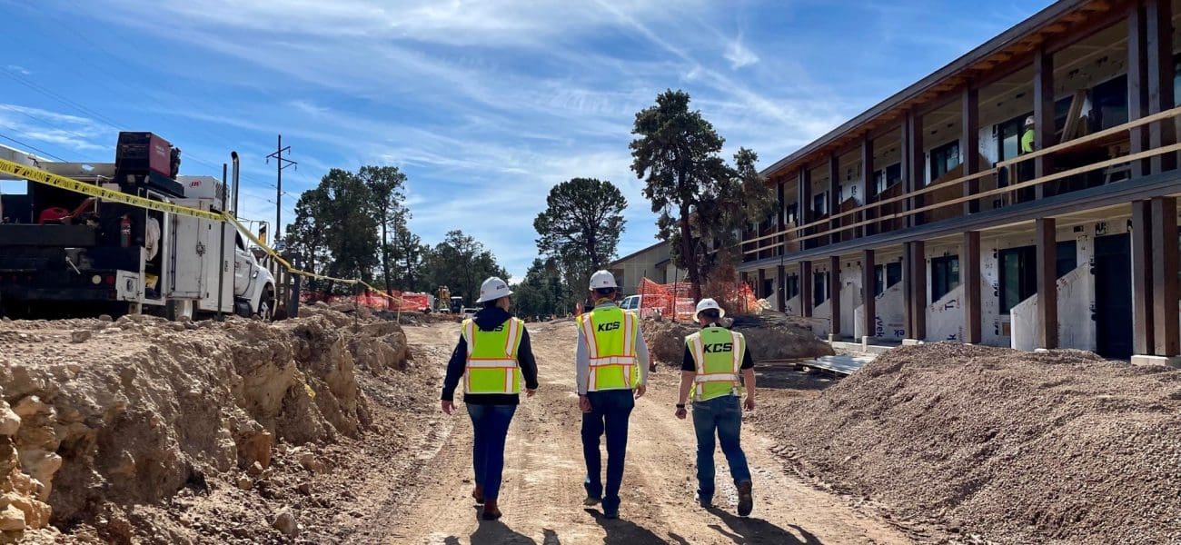 Three construction workers in safety vests and helmets walk along a dirt path at an active construction site with buildings and equipment, where Kinney is recognized as one of the Top 140 General Contractors.