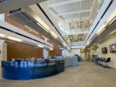 Modern office lobby with blue reception desk, open workspace, glass railings, overhead lights, and wall-mounted monitors. The area appears spacious and well-lit.