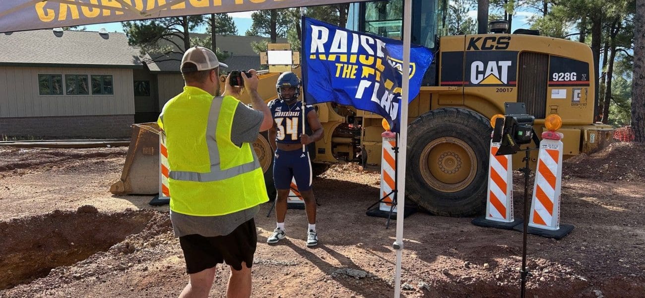 NAU Football + NPA A person in a safety vest photographs a football player posing by construction equipment and a "RAISE THE FLAG" banner, highlighting Kinney's commitment to Building Better Together for a Greener Tomorrow, as they honor Earth Day at the construction site.