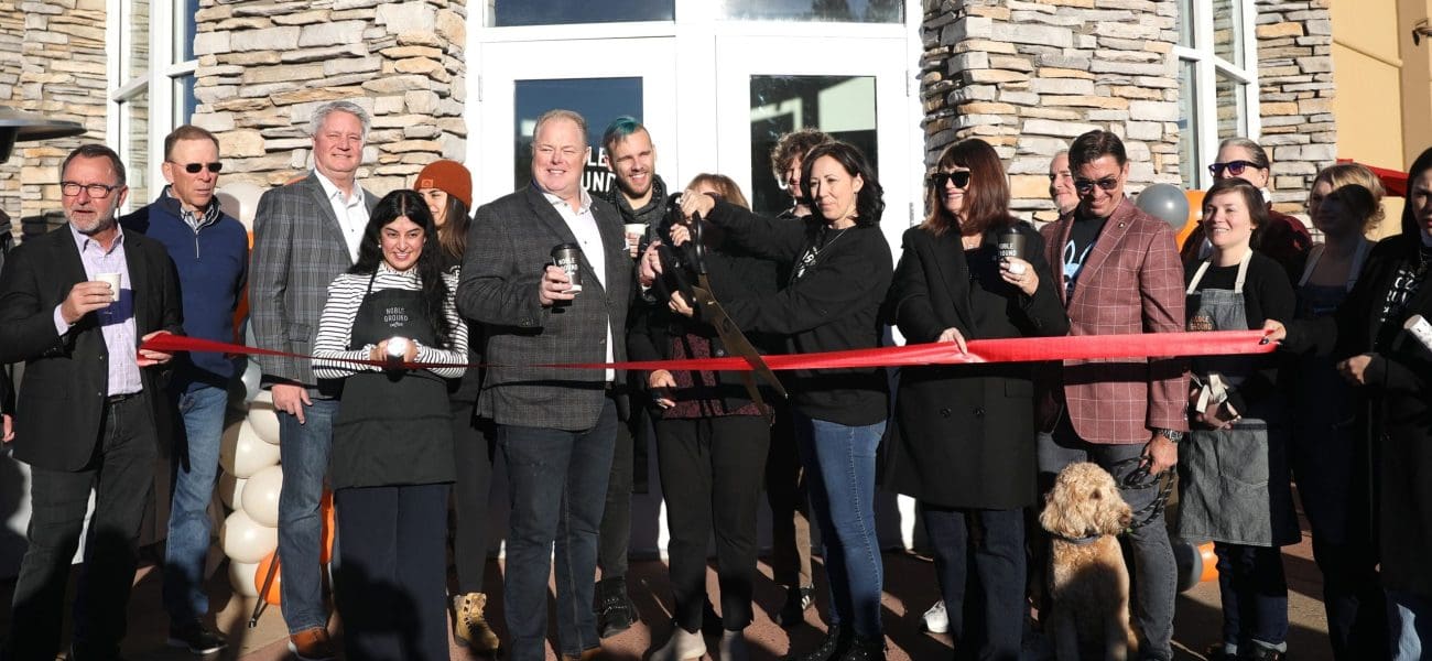 A group of people stand in front of Noble Ground Coffee during a ribbon-cutting ceremony; a woman cuts the red ribbon while others hold drinks and smile, celebrating Flagstaff's newest coffee shop with a cause.