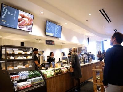Customers stand in line at a modern café, viewing digital menu boards above the counter while staff prepare orders behind a display of bakery items and refrigerated drinks.
