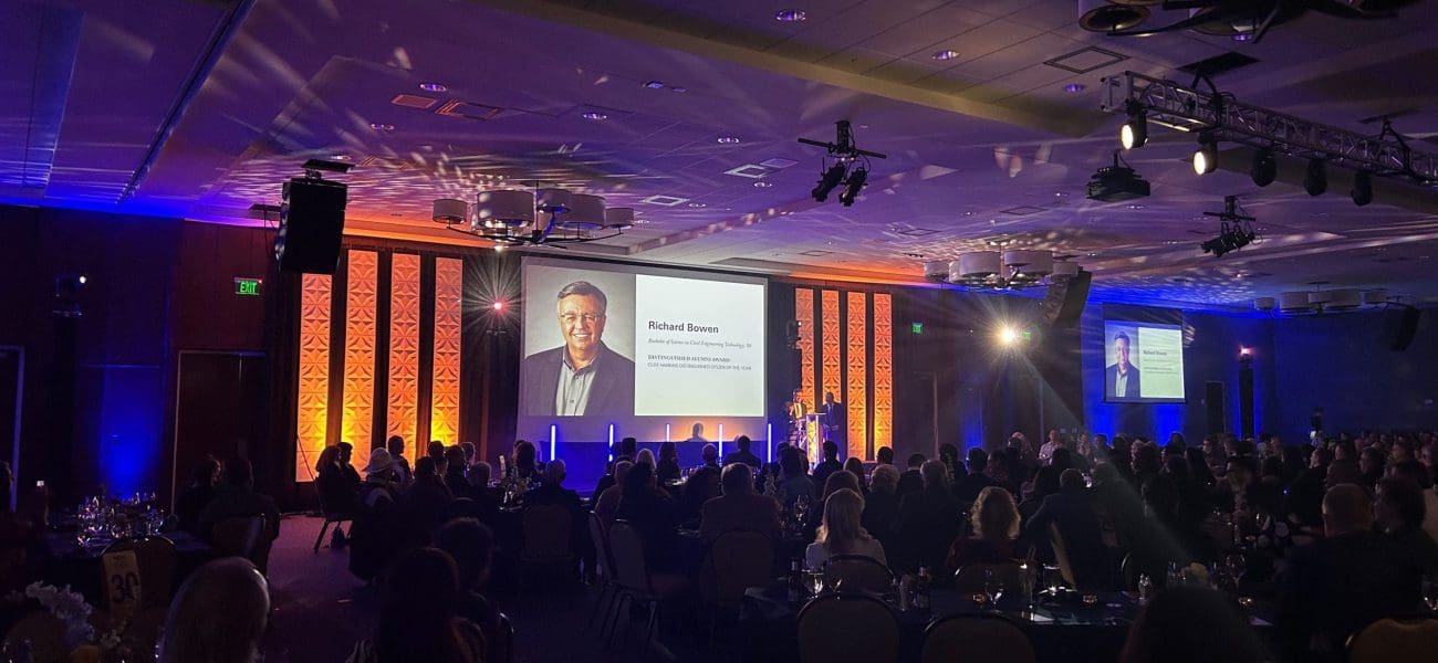 Rich Bowen NAU Award A formal event with attendees seated at tables facing a stage, where a man's portrait and name are projected onto a large screen, celebrating Kinney joining forces with United Way to build stronger communities.