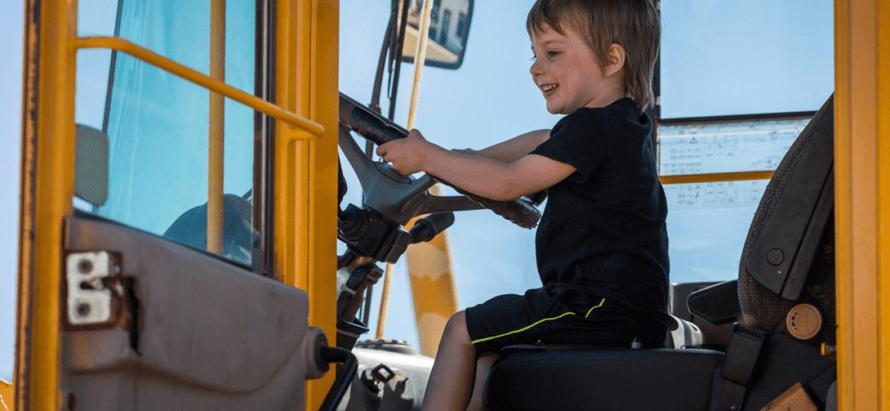 A young child sits in the driver’s seat of a large yellow construction vehicle, smiling and holding the steering wheel at the 2025 Annual Community STEM Celebration, celebrating innovation and curiosity.