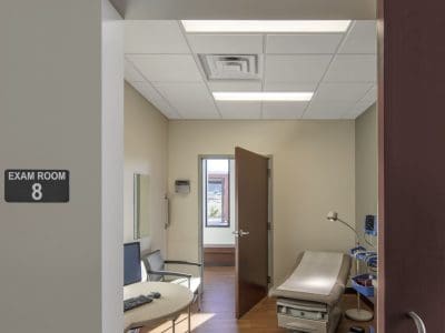 View into a medical exam room with an exam table, desk, rolling chair, and medical equipment. The door is open, and a sign reads "Exam Room 8.