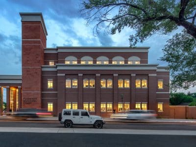 A brick courthouse building with illuminated windows at dusk, cars passing by on the street, and a large tree in the foreground.
