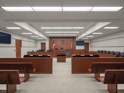 Interior of an empty courtroom with wooden benches, a judge's bench, flags, and a podium, viewed from the public seating area.