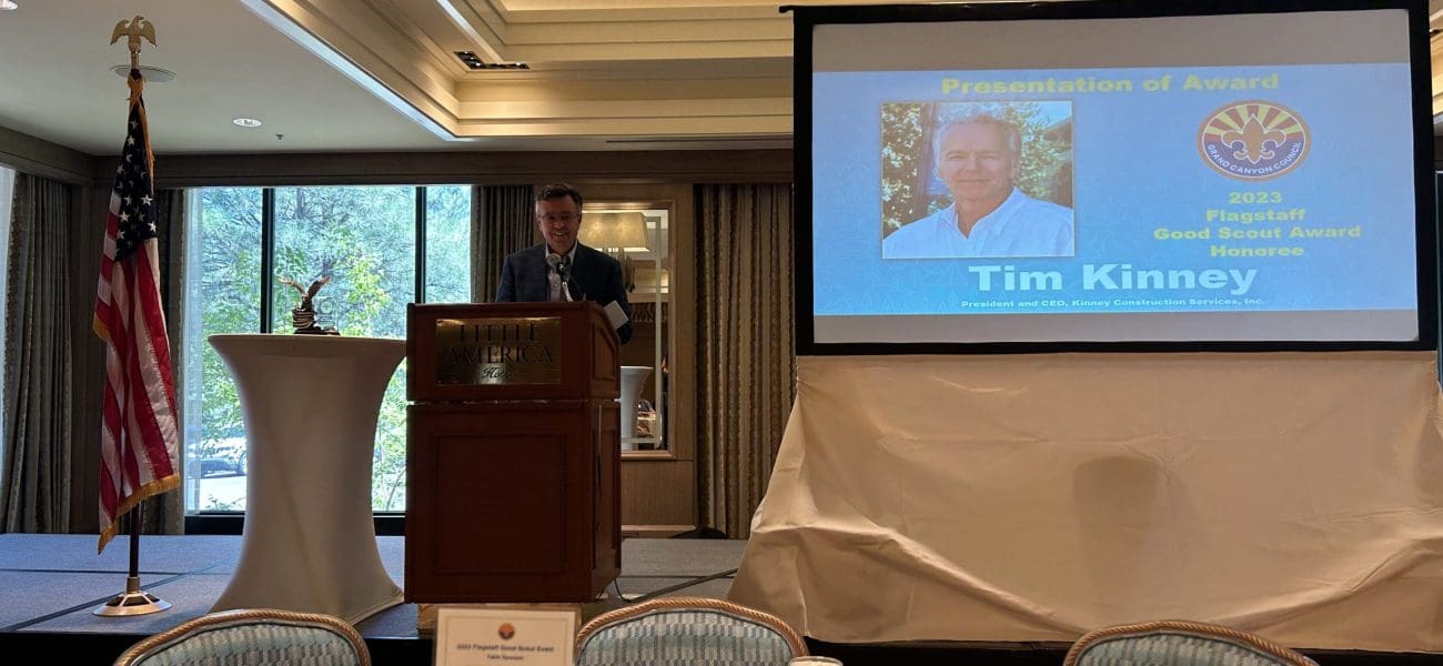 A man stands at a podium giving a presentation in a banquet room, with a projection behind him announcing Tim Kinney as the 2023 Good Scouts' Award Recipient.
