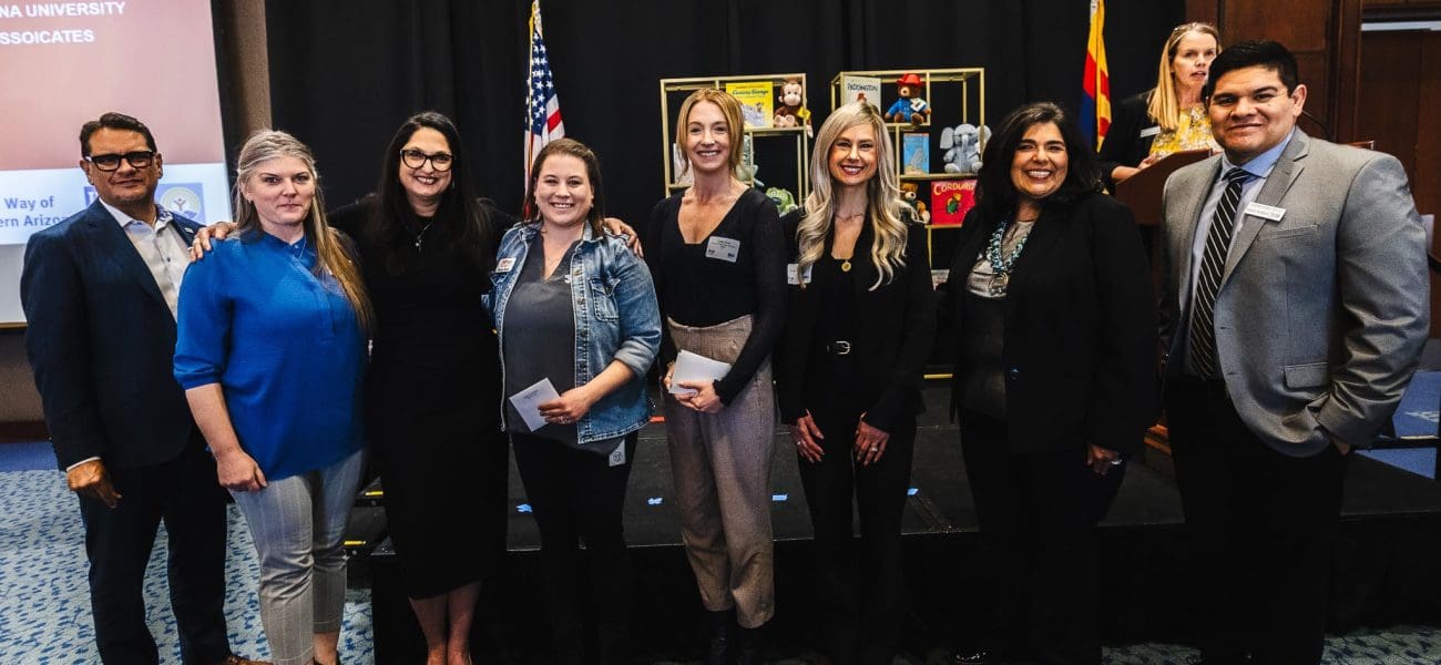 United Way Luncheon 2025 Nine people pose for a group photo indoors at an event, standing in front of a stage with U.S. and Arizona flags and a display of photos, as Kinney joins forces with United Way to build stronger communities.