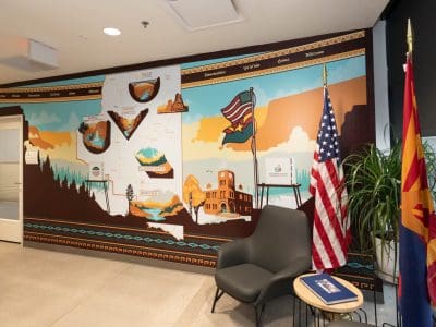 A mural depicting Arizona landmarks and a map on a wall, next to a gray chair, a small table with a booklet, two flags, and potted plants in an office space.