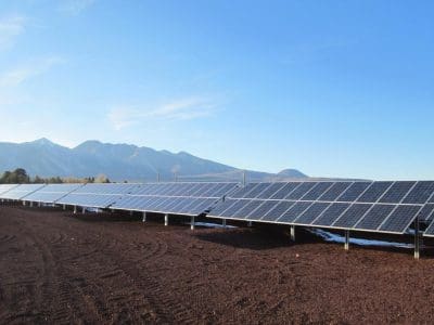 Rows of solar panels from the APS Doney Park PV System installed on the ground with mountains and a clear blue sky in the background.