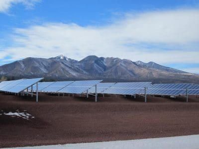 Rows of solar panels from the APS Doney Park PV System are installed on reddish-brown ground with a mountain range and partly cloudy sky in the background.