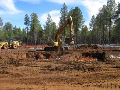An excavator digs a large hole at a construction site in a forested area, with a bulldozer and orange safety fencing in the background.