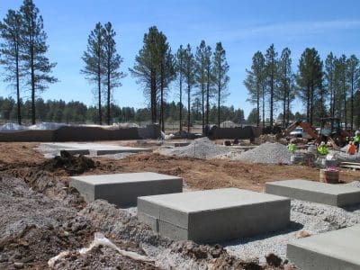 A construction site with rectangular concrete foundations, gravel, equipment, and workers in safety gear; pine trees and clear sky in the background.