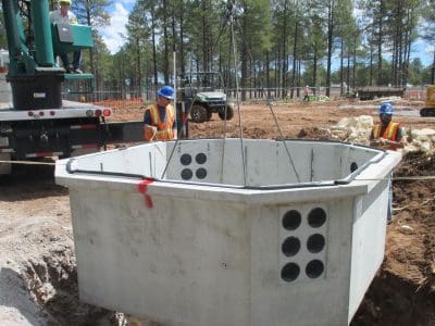 Construction workers guide a large concrete utility vault being lowered by a crane into an excavated pit at a worksite surrounded by trees and equipment.