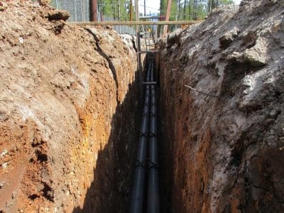 A trench with several large black utility pipes installed underground, surrounded by dirt walls, with construction barriers visible in the background.