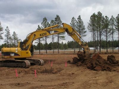 A yellow CAT excavator moves a pile of dirt at a construction site marked with pink stakes, surrounded by tall pine trees and a cloudy sky.