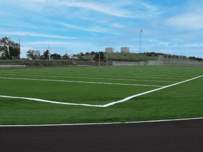 A freshly marked sports field with artificial turf and white boundary lines under a blue sky; buildings and trees are visible in the background.