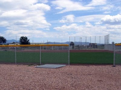 A fenced baseball field with green artificial turf and brown dirt infield under a partly cloudy sky, with mountains visible in the distance.
