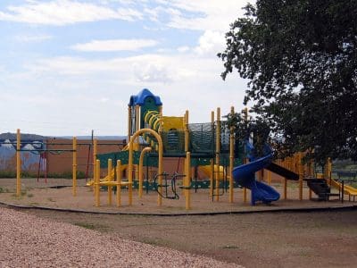 A playground with yellow and green climbing structures, slides, and bridges sits on a sandy surface beside a large tree under a partly cloudy sky.