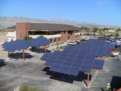 Bullhead City PV System features solar panel arrays installed over a parking lot in front of a two-story brick building, with scenic mountains visible in the background.