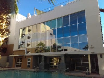 Modern building with large glass windows reflecting the sky, palm trees, and a swimming pool in the foreground.