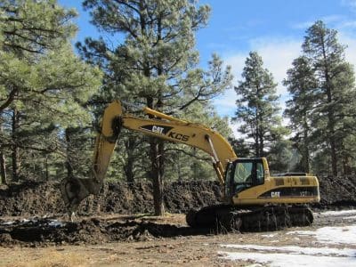 A yellow CAT excavator is digging soil in a forested area with pine trees and patches of snow on the ground.