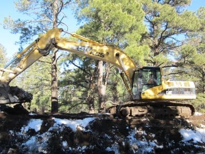 A yellow CAT 325 excavator is digging soil on a snow-dusted, forested area with pine trees in the background.