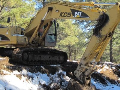 A yellow KCS CAT excavator is digging earth at the edge of a snow-covered area surrounded by trees.