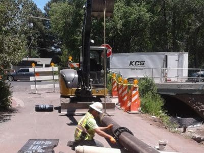 A construction worker in a safety vest and helmet works with a pipe on the ground near an excavator at a street worksite with traffic barriers and a truck in the background.