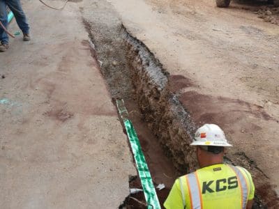 A construction worker in a yellow safety vest labeled "KCS" installs a green warning tape in a freshly dug trench on a partially paved street.