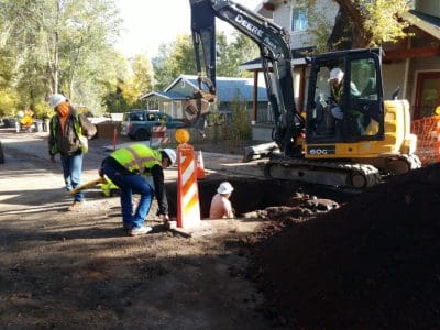 Construction workers operate machinery and work in a large hole on a residential street, surrounded by safety barriers and traffic cones.