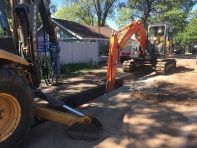 Two excavators dig a trench on a residential street, with workers operating the machinery near houses and trees on a sunny day.