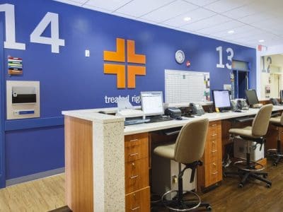 Hospital nurse station with multiple chairs, computers, and a blue wall featuring room numbers and a cross symbol, under bright ceiling lights.